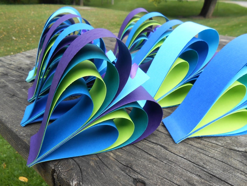 Pu&ograve; includere: Un gruppo di cuori di carta fatti di carta blu, verde e viola. I cuori sono piegati in modo da creare un effetto a strati.