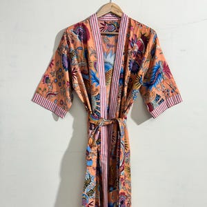 May include: A peach-colored, floral-print robe with a matching belt. The robe features a vibrant pattern of blue, purple, and red flowers and leaves. The edges are trimmed with pink stripes. The robe is hanging on a wooden hanger.