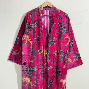 May include: A vibrant pink robe with a jungle-themed pattern featuring zebras, leopards, flamingos, and tropical plants. The robe has a striped lining and a shawl collar. The robe is hanging on a wooden hanger.