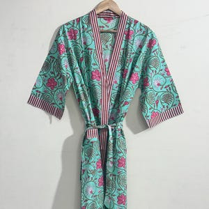 May include: A turquoise floral print robe with pink flowers and brown leaves. The robe has a striped trim in burgundy and white along the edges and a matching belt. The robe is hanging on a wooden hanger.