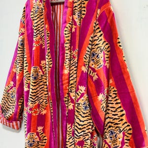 May include: A long-sleeved robe featuring a vibrant pattern of orange and black tigers set against vertical stripes of bright magenta and orange. The garment includes a solid magenta trim along the front opening and cuffs with a white and orange striped lining.
