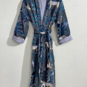 May include: A gray robe featuring a jungle-themed print with leopards, toucans, and palm leaves in shades of blue, white, and yellow. The garment includes long sleeves with folded cuffs, a tie belt at the waist, and a white and pink striped lining.