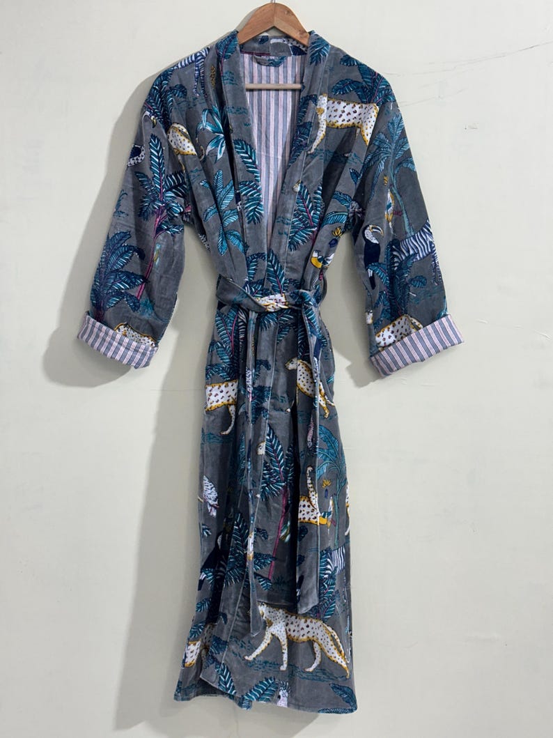May include: A gray robe featuring a jungle-themed print with leopards, toucans, and palm leaves in shades of blue, white, and yellow. The garment includes long sleeves with folded cuffs, a tie belt at the waist, and a white and pink striped lining.