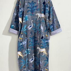 May include: A long-sleeved gray robe featuring a repeating pattern of leopards, zebras, flamingos, toucans, and tropical palm trees. The garment has a V-neckline and cuffs with a white and gray striped lining, hanging from a wooden hanger.