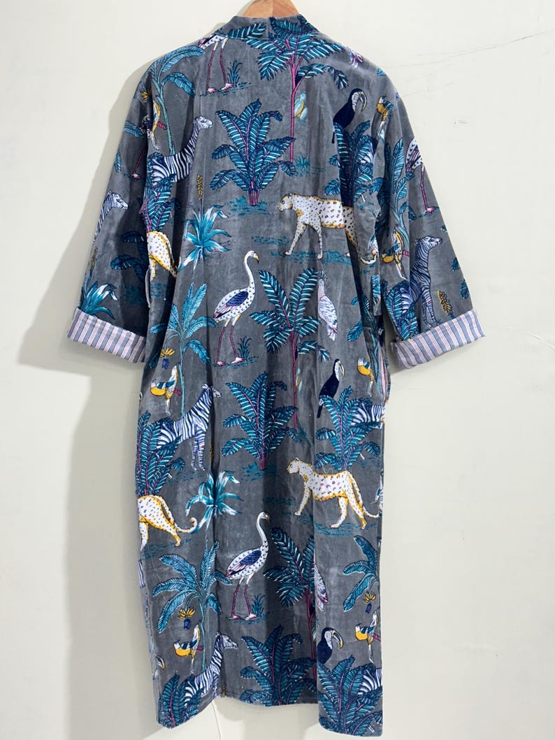 May include: A long-sleeved gray robe featuring a repeating pattern of leopards, zebras, flamingos, toucans, and tropical palm trees. The garment has a V-neckline and cuffs with a white and gray striped lining, hanging from a wooden hanger.