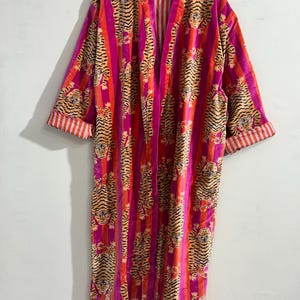 May include: A long-sleeved robe featuring a vibrant pattern of tigers interspersed with vertical stripes in shades of bright pink and orange. The garment has an open front design and folded cuffs that reveal a white and orange striped interior lining.