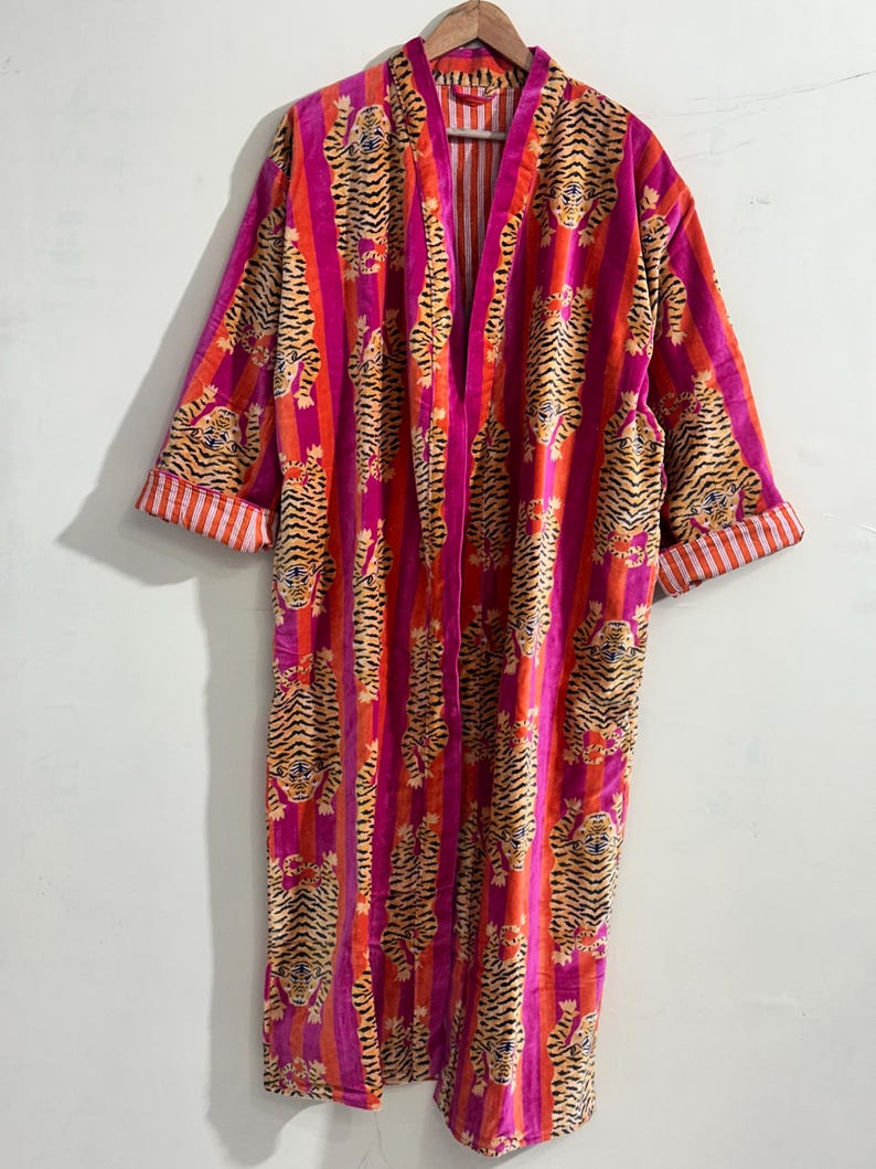 May include: A long-sleeved robe featuring a vibrant pattern of tigers interspersed with vertical stripes in shades of bright pink and orange. The garment has an open front design and folded cuffs that reveal a white and orange striped interior lining.