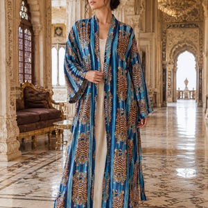 May include: A floor-length open-front kimono robe featuring a repeating tiger pattern in shades of orange, black, and white, set against vertical blue and light blue stripes. The garment is worn over a simple, solid cream-coloured sleeveless dress.