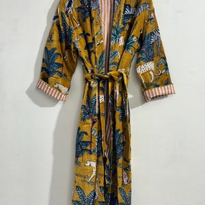 May include: A mustard yellow robe featuring a repeating print of zebras, leopards, and palm trees in shades of blue, white, and black. The garment includes long sleeves with white and pink striped cuffs, a matching tie belt, and an open front design.