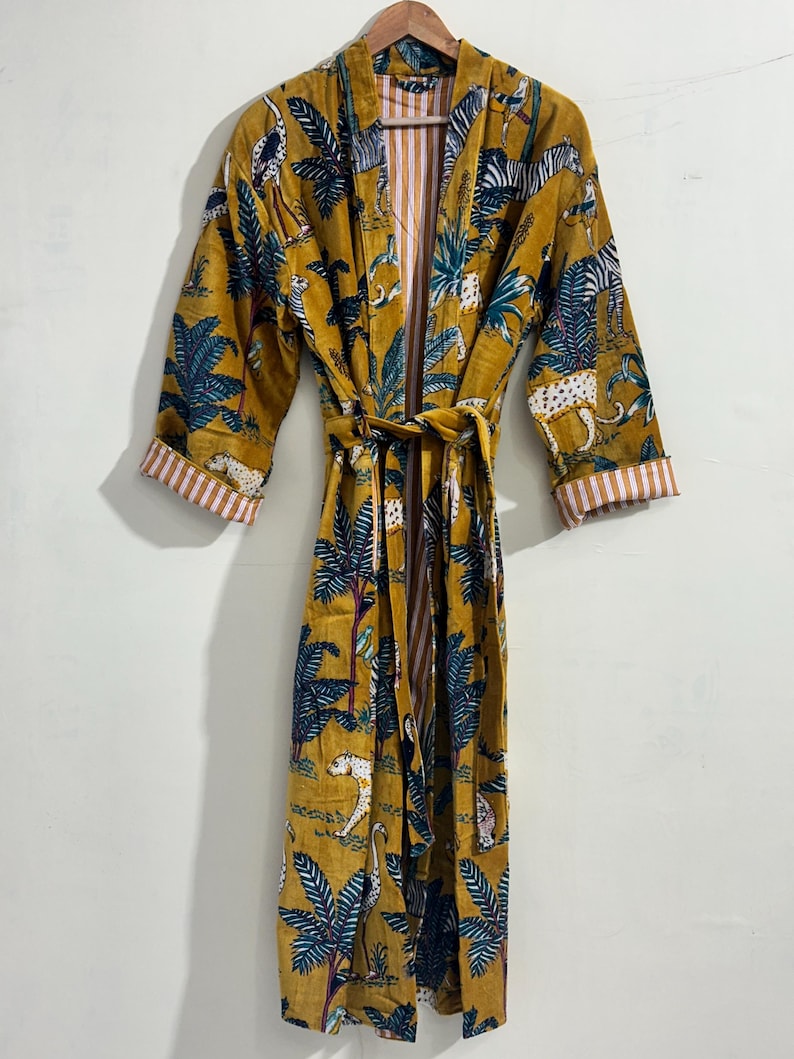 May include: A mustard yellow robe featuring a repeating print of zebras, leopards, and palm trees in shades of blue, white, and black. The garment includes long sleeves with white and pink striped cuffs, a matching tie belt, and an open front design.