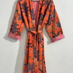 May include: A long-sleeved orange robe featuring a repeating print of leopards, zebras, and palm trees. The garment includes a matching tie belt at the waist and cuffs that reveal a white and red vertical striped lining on the interior.