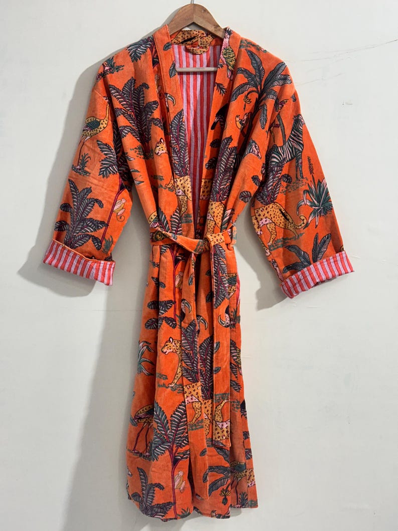 May include: A long-sleeved orange robe featuring a repeating print of leopards, zebras, and palm trees. The garment includes a matching tie belt at the waist and cuffs that reveal a white and red vertical striped lining on the interior.