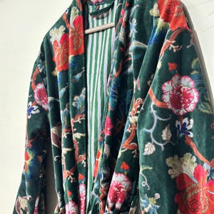 May include: A dark green velvet robe featuring a vibrant floral pattern with red, pink, and blue blossoms on winding branches. The garment includes a matching tie belt and a contrasting interior lining with thin vertical white and green stripes.