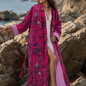Puede incluir: Una bata larga abierta de terciopelo fucsia vibrante con un estampado detallado de flores y pájaros. La prenda incluye mangas largas con puños a rayas blancas y fucsias en contraste y un forro a rayas a juego visible en el borde delantero.