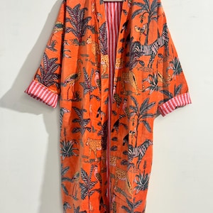 May include: A long, open-front robe in a vibrant orange hue featuring a repeating print of jungle animals, including zebras, leopards, and flamingos, amidst palm trees. The garment has wide sleeves with cuffs that reveal a red and white striped lining.