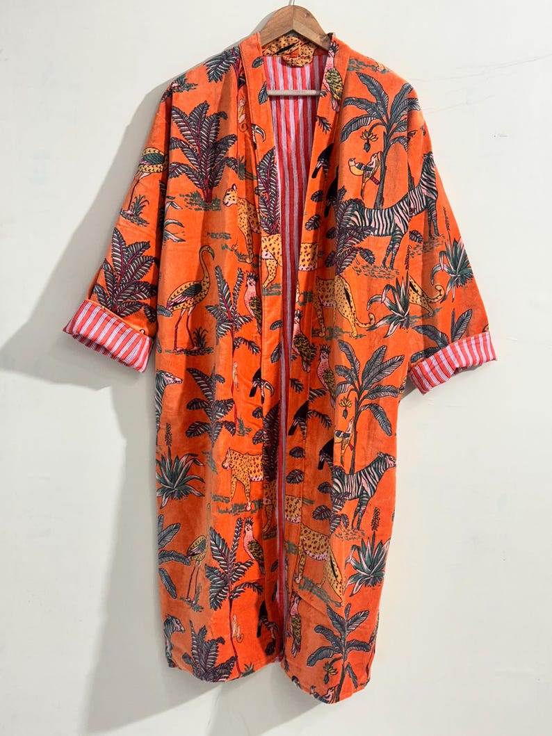 May include: A long, open-front robe in a vibrant orange hue featuring a repeating print of jungle animals, including zebras, leopards, and flamingos, amidst palm trees. The garment has wide sleeves with cuffs that reveal a red and white striped lining.