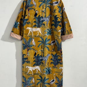 May include: A mustard yellow robe featuring a repeating jungle print with leopards, zebras, toucans, and cranes among palm trees. The garment has long sleeves with white and orange striped cuffs and is displayed on a wooden hanger.