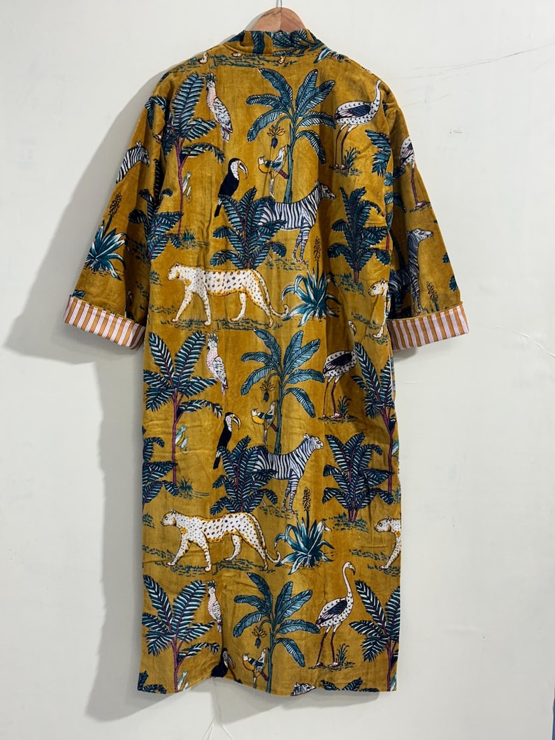 May include: A mustard yellow robe featuring a repeating jungle print with leopards, zebras, toucans, and cranes among palm trees. The garment has long sleeves with white and orange striped cuffs and is displayed on a wooden hanger.
