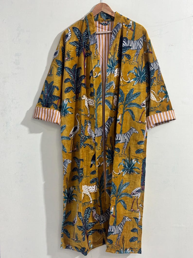 May include: A long-sleeved mustard yellow robe featuring an all-over print of zebras, leopards, and tropical palm trees. The garment has an open front design with white and orange vertical striped cuffs and an interior lining in the same pattern.