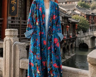 May include: A long, open-front velvet duster coat in a vibrant blue hue, featuring a colorful floral and bird pattern. The garment has long sleeves with white and blue striped cuffs, a white inner lining, and a relaxed, floor-length silhouette.