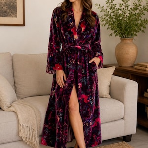 May include: A floor-length velvet robe in a deep plum purple hue, featuring a vibrant floral pattern in shades of bright pink, red, and green. The robe includes long sleeves, a shawl collar, a matching tie belt at the waist, and two side pockets.