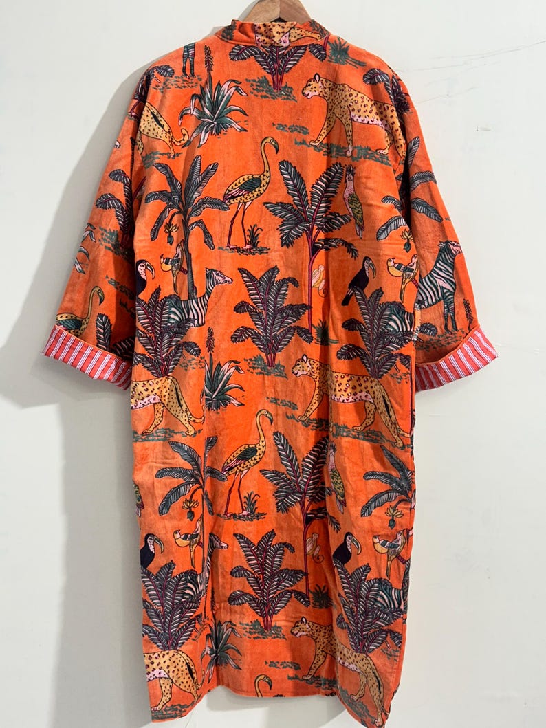 May include: An orange long-sleeve robe featuring a repeating print of leopards, flamingos, toucans, and zebras among palm trees. The garment has a mandarin collar and cuffs with a red and white vertical striped pattern on the interior lining.