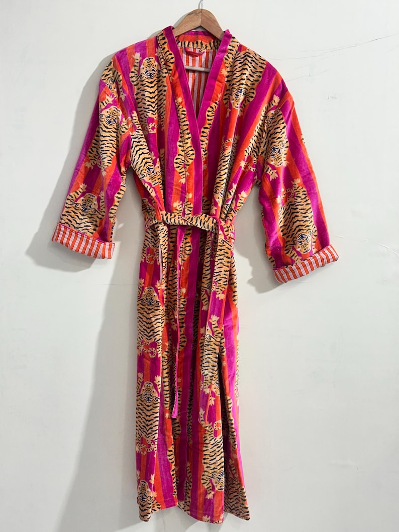 May include: A long-sleeved robe featuring a vibrant pattern of orange and black tiger faces set against vertical stripes of bright pink and orange. The garment includes a matching tie belt at the waist and folded cuffs with a white and orange striped lining.