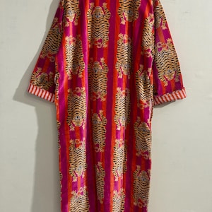 May include: A long-sleeved robe featuring a repeating tiger pattern over vertical stripes of bright pink and orange. The garment has a mandarin-style collar and cuffs with a white and orange striped trim, hanging from a wooden hanger.
