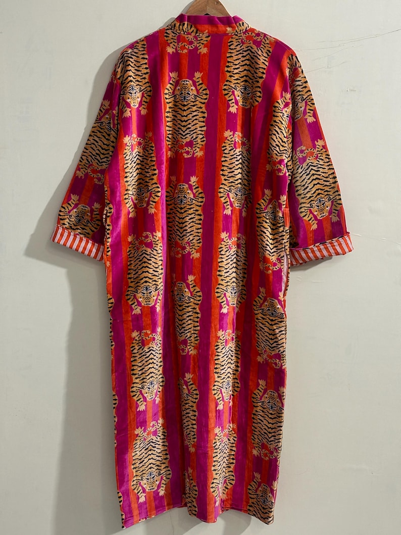 May include: A long-sleeved robe featuring a repeating tiger pattern over vertical stripes of bright pink and orange. The garment has a mandarin-style collar and cuffs with a white and orange striped trim, hanging from a wooden hanger.
