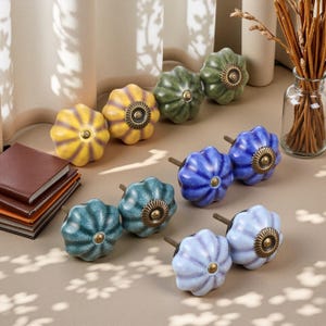 May include: A collection of ceramic cabinet knobs in various colors, including yellow, green, blue, and light blue. Each knob has a flower-like design with a brass-colored center and a metal stem for installation. A stack of brown notebooks is in the background.