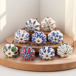 May include: A collection of ten ceramic cabinet knobs with floral designs. The knobs are white with blue, red, and green painted patterns. Each knob has a brass-colored stem and is arranged on a wooden display.