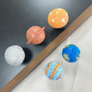 May include: A collection of colorful planet-shaped decorative objects. The planets include a textured silver sphere, an orange sphere with white spots, a brown sphere with a ring, a blue and green sphere, and a sphere with blue and orange stripes.