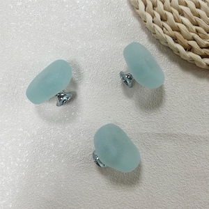 May include: Three seafoam green cabinet knobs with a frosted finish and silver-toned bases are arranged on a textured white surface. The knobs have an oval shape and are evenly spaced, suggesting a set for furniture.