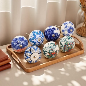 Puede incluir: Una colección de ocho pomos de cerámica decorativos con diseños florales en azul, blanco y verde. Los pomos se exhiben en una bandeja de madera, mostrando sus intrincados patrones y herrajes metálicos.