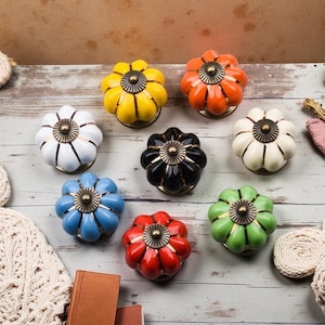 May include: A selection of colourful ceramic cabinet knobs in shades of yellow, orange, white, black, blue, red, and green. Each knob features a flower-like design with a brass-coloured centre and base. These decorative knobs are perfect for adding a splash of colour to furniture.
