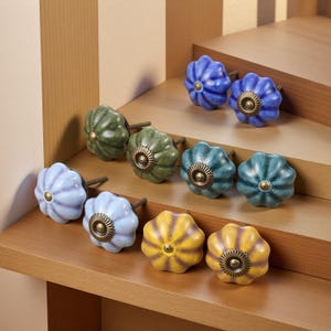 Op de afbeelding: Een verzameling decoratieve keramische kastknoppen in verschillende kleuren, waaronder blauw, groen en geel. Elke knop heeft een bloemachtige vorm met een messingkleurig midden en steel. De knoppen zijn gerangschikt op houten planken.