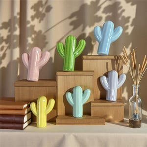 May include: A selection of ceramic cactus ornaments in pastel shades of pink, green, blue, yellow, and lilac. The cacti are displayed on wooden blocks of different heights, creating a decorative arrangement. A clear glass bottle with reeds is on the right.