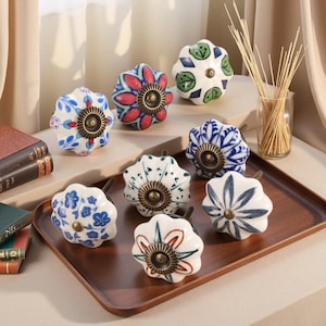 May include: Eight ceramic cabinet knobs with floral patterns in blue, red, green, and white. Each knob has a brass-coloured base and stem. The knobs are displayed on a wooden tray.