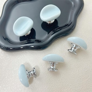 May include: Light blue cabinet knobs with a polished silver base. The knobs have a smooth, oval shape. Several knobs are arranged on a black, wavy-edged tray, with others scattered on a neutral background.