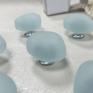 May include: A collection of pale blue, sea glass-style cabinet knobs. Each knob has a rounded, organic shape and is mounted on a silver-toned base. The knobs are arranged on a textured, off-white surface.