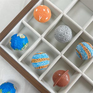 May include: A collection of planet-themed decorative spheres in a wooden tray. The spheres feature detailed designs representing Earth, the Moon, and other planets, with vibrant colors like blue, orange, and silver. The tray has multiple compartments.
