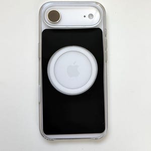 May include: A clear smartphone case featuring a black rectangular insert on the back. The case includes a circular white magnetic charging attachment centered on the black insert, which displays a light gray Apple logo in the middle.
