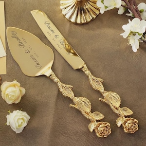 Puede incluir: Un juego de cuchillo y servidor de pastel dorado con detalles florales y de hojas. El juego está grabado con "Destin & Bradley" y "OCT. 03. 2025". Los mangos presentan diseños en forma de rosa.