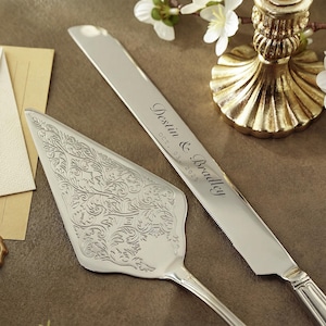 Puede incluir: Juego de cuchillo y servidor de pastel plateado. El cuchillo está grabado con "Destin & Bradley" y "OCT. 03. 2025". El servidor tiene un diseño floral ornamentado. Un candelabro dorado y flores blancas están en el fondo.