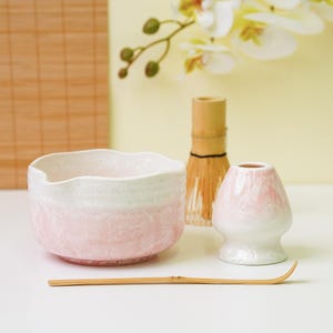 Può includere: Un set da tè matcha con una ciotola in ceramica rosa e bianca, un frullino in bambù, un portafrullino in ceramica rosa e bianca e un cucchiaio in bambù. Per preparare il tè matcha.