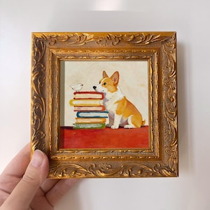 May include: A framed painting featuring a corgi dog sitting next to a stack of colorful books. A small white bird is perched on top of the books. The frame is ornate with a gold finish.
