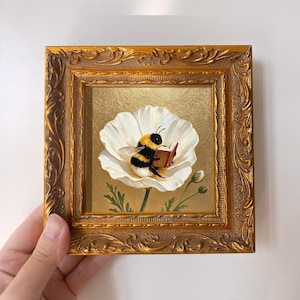 以下が含まれることがあります： 装飾が施された金色のフレームに入った正方形の絵画。黒と黄色の縞模様のマルハナバチが大きな白い花びらの中に座り、メタリックゴールドの背景の前で小さな茶色の本を持って読んでいる様子が描かれています。