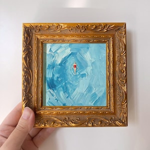 May include: A small square oil painting featuring a person in a red swimsuit floating in textured blue water, encased in an ornate, antique-style gold frame with intricate carved detailing along the edges and a polished metallic finish.