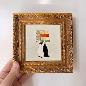 Puede incluir: Una obra de arte enmarcada que representa un pingüino llevando una pila de libros coloridos sobre su cabeza. El pingüino es blanco y negro con pies rojos. El marco es dorado, ornamentado y cuadrado.