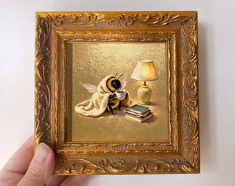 May include: A small square painting in an ornate gold frame depicting a bee wrapped in a cream-colored blanket, holding a steaming white cup. Beside the bee is a small lit table lamp and a stack of three books on a gold-toned surface.